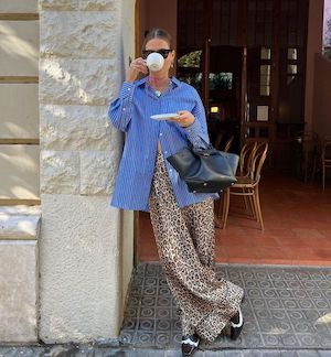 brunette woman wearing sunglasses, blue long sleeve button up, black purse, and cheetah pants sipping coffee leaning against a pillar