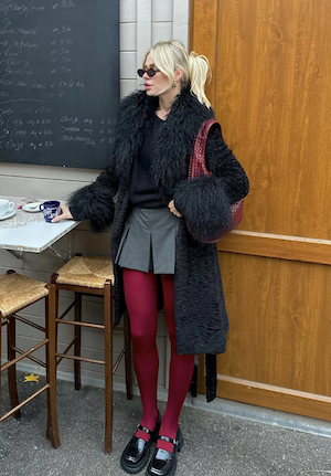 a blonde woman wearing long black fur coat, grey mini skirt, and red tights standing next to a table outside