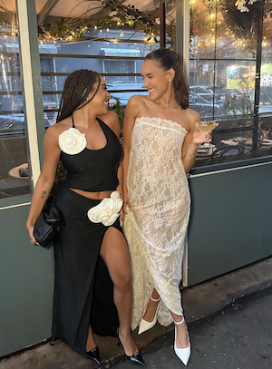 two dark-haired girls smiling at each other, one wearing a white lace maxi dress and white heels; the other wearing a black halter top and black long skirt with a slit, and white roses on both top and bottom