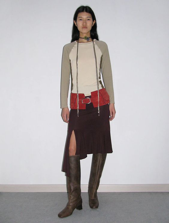 a dark-haired woman standing against a wall wearing a white and grey long sleeve, red belt, brown midi skirt, tall dark boots, and a string choker necklace