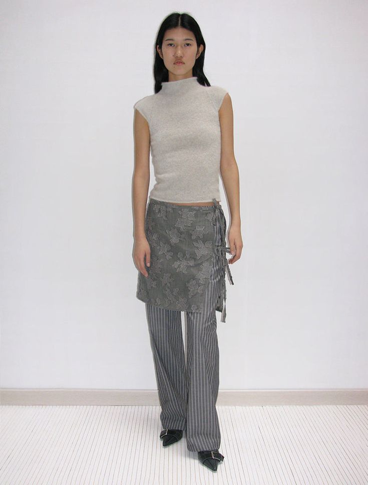 dark-haired woman standing against wall wearing beige top and gray wrap skirt over gray pants