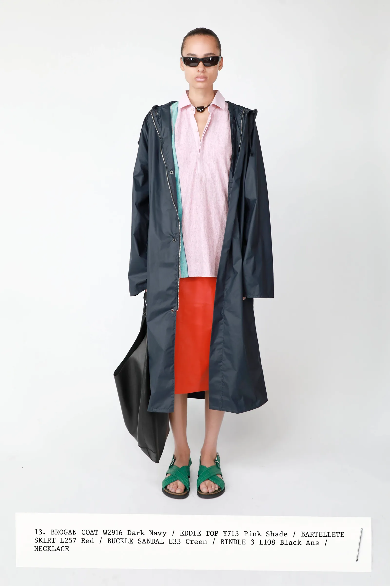 model standing against white backdrop wearing a black trench coat, pink polo top, orange midi skirt, green sandals, and black sunglasses