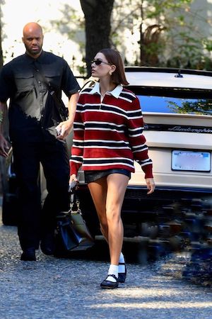 Hailey Bieber walking on street in red and black striped long sleeve polo, black mini skirt, socks and black ballet flats