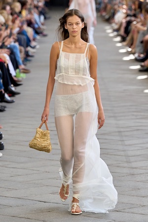 brunette model wearing sheer white maxi dress holding a yellow handbag on the runway