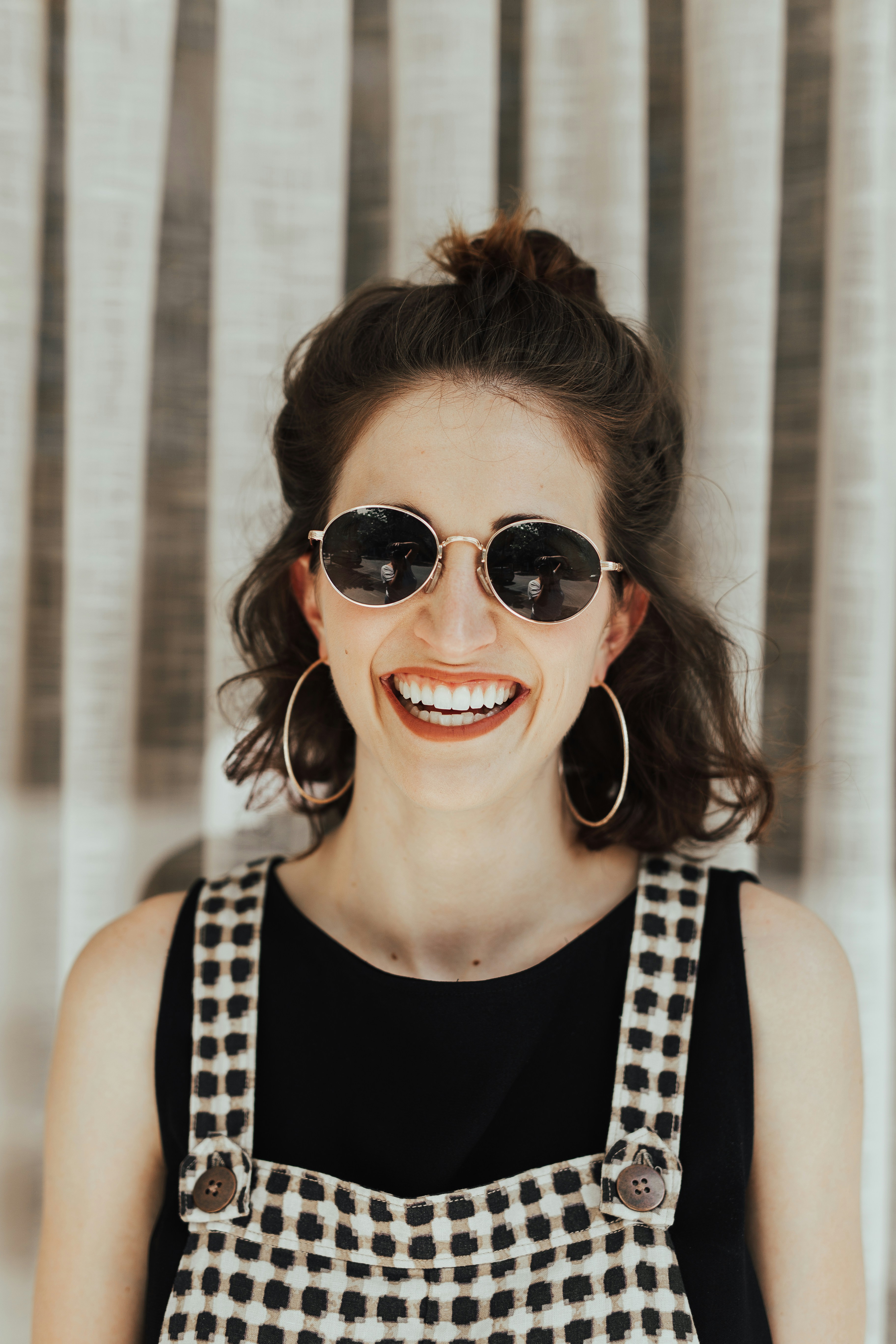 headshot of a smiling woman with short brunette hair wearing dark sunglasses and checkered overalls